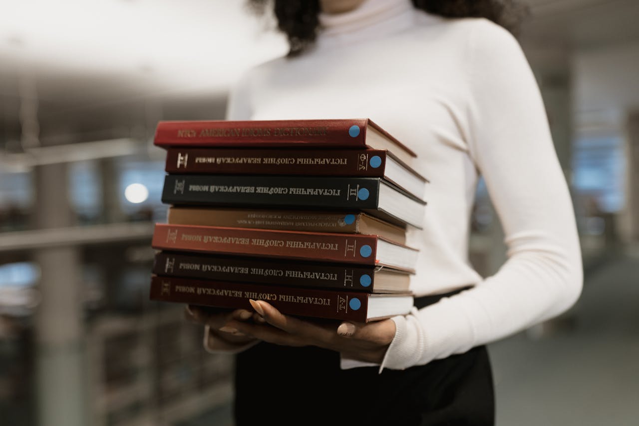 A woman holds a stack of academic books in a library setting, emphasizing education.